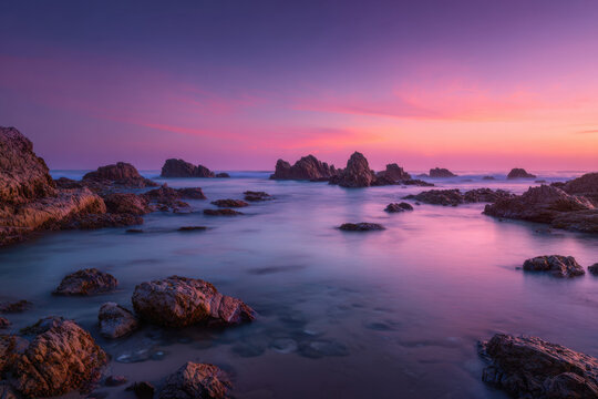 Dreamy coastal seascape at sunset with pink and purple sky, rocky shoreline and smooth water creating serene minimalist atmosphere