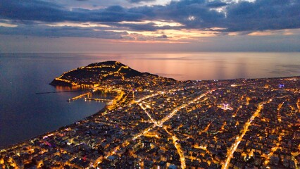 A drone-captured dusk panorama of Alanya, Turkey, showing the illuminated city, peninsula and harbor against a calm sea and twilight sky