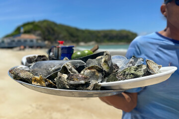 Close-up view of fresh oysters served on a tray with crushed ice and whole limes, held by a vendor on a sunny beach.