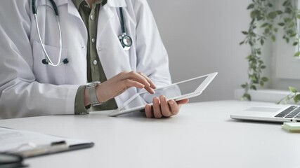 Female doctor in lab coat and stethoscope using digital tablet at desk in medical office, accessing online patient information. Medicine and health care concept - Powered by Adobe