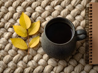 A comforting cup of dark coffee with yellow autumn leaves and a spiral notebook rests on a chunky knit blanket creating cozy mood.