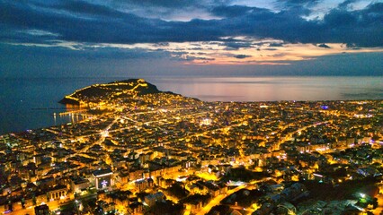 A drone-captured dusk panorama of Alanya, Turkey, showing the illuminated city, peninsula and harbor against a calm sea and twilight sky