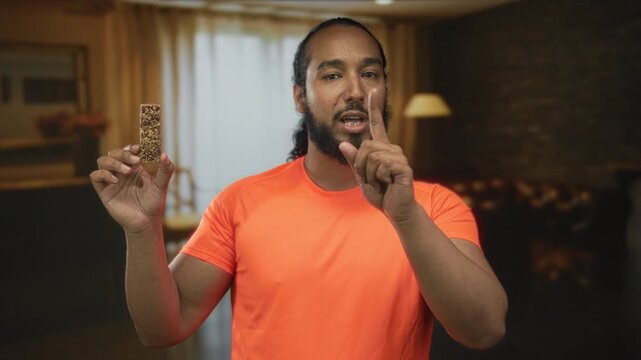Man holding granola bar and points finger to snack in a building living room; nutrition tip confidence.