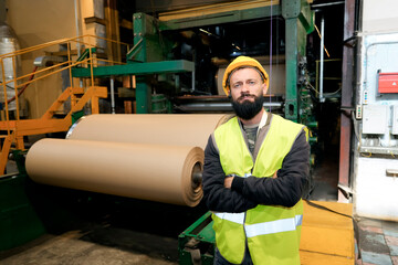 In a recycling plant, a man with a beard wearing a helmet and safety vest stands near a machine. He...