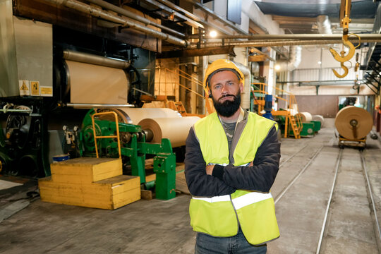A man with a beard stands confidently in a recycling factory, surrounded by machinery and large cardboard rolls. He wears a safety helmet and a reflective vest, ensuring safety in the busy workshop