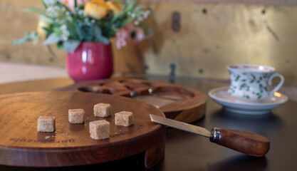 Close-up of brown sugar cubes and a small wooden knife on a round cutting board in a cozy cafe setting. Tea or coffee cup and flowers are blurred in the background. Breakfast concept