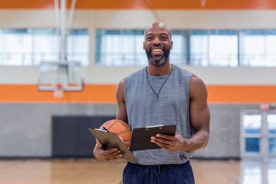 Smiling basketball coach holding clipboard and ball in gymnasium setting