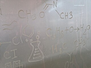 An abstract, close-up view of various chemical formulas, molecular structures (including a benzene ring), and a conical flask symbol lightly drawn or scratched onto a foggy or frosted glass surface.