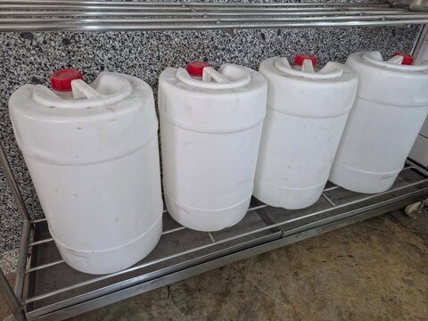 Four identical, large white plastic jerrycans or drums with red caps, containing industrial or chemical liquids, lined up on a stainless steel shelf against a granite-patterned wall.
