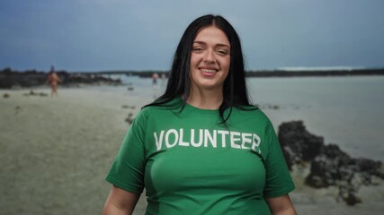 Woman smiling at seaside beach in green volunteer shirt, showing excitement and enthusiasm, with ocean waves in the background under a clear blue sky.