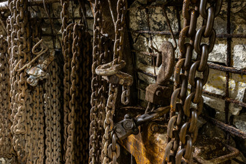 Heavy tools and equipment inside an underground mine, with steel chains, pneumatic hoses, and supply lines in active use. The scene shows industrial work conditions, mining technology, and harsh subte