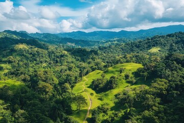 An aerial view shows a lush green landscape with rolling hills and a winding path through the trees under a blue sky with white clouds.