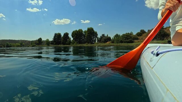 Young woman paddles inflatable boat on tranquil lake with an orange oar on beautiful summer day, enjoy serene landscape and reflection of clouds on water's surface