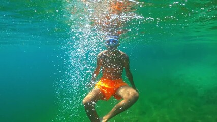 Young boy wearing swimming trunks and snorkel mask diving into clear turquoise sea on sunny summer day. Teenager boy descent creates bubbles and ripples on water's surface