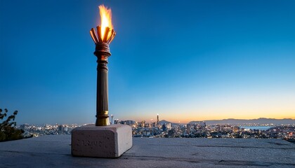 a lit torch stands on a stone pedestal with a blurred city skyline in the background under a clear blue sky
