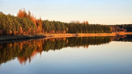 Tranquil autumn forest reflected in calm lake waters at sunset. Serene landscape with vibrant trees, natural symmetry and warm evening light