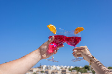 Hands toasting with red cocktails and orange slices under clear blue sky