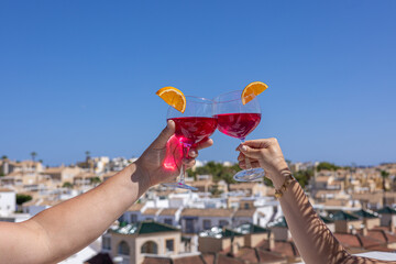 Cheers with red cocktails above Mediterranean rooftops under blue sky