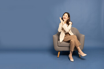 Young beautiful Asian businesswoman surprise and delight and sitting on armchair isolated over blue background, Thrilled excited concept