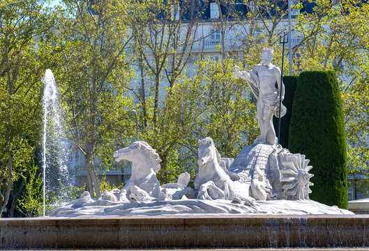 Close-up of Madrid&rsquo;s Fuente de Neptuno, showing the white marble Neptune with trident, surrounded by horses and a strong water jet, all framed by vibrant green trees