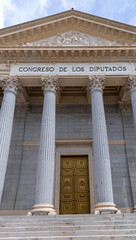 The neoclassical facade of the Spanish Congress of Deputies features six columns, a pediment, steps, and two large bronze lion statues flanking the main golden doorway