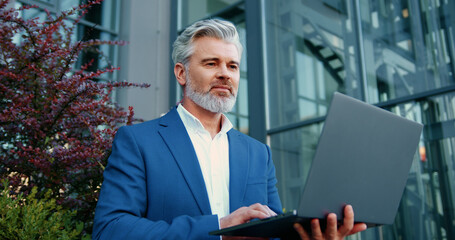 Mature businessman working thoughtfully on a laptop outdoors with a modern glass building in the background.