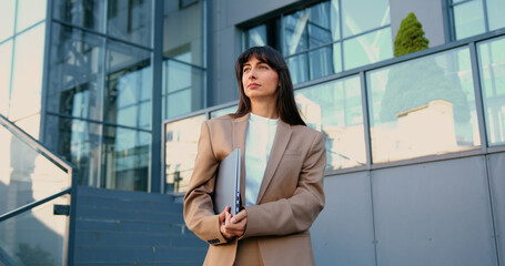 Confident young woman in business suit holding laptop, standing in front of modern office building, smiling softly as she looks into the distance.