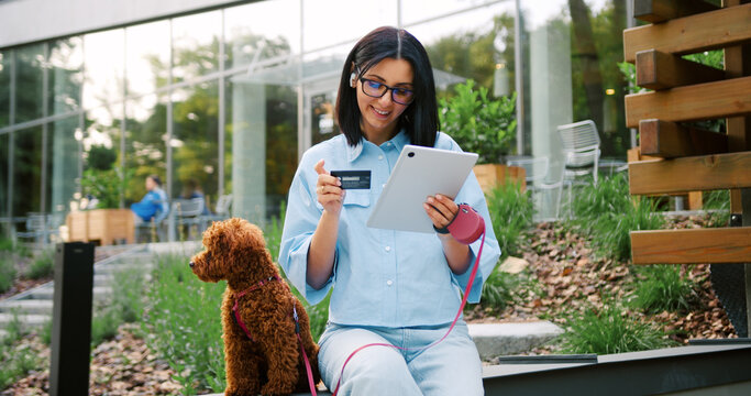 Young woman in glasses wearing casual clothes entering credit card information for an online purchase on a tablet while her Maltipoo dog sits near on a leash nearby in a green city park