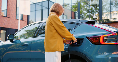 Fototapeta premium Modern stylish woman connects charging cable parked electric vehicle. Concept of eco transport, green energy, and modern lifestyle.