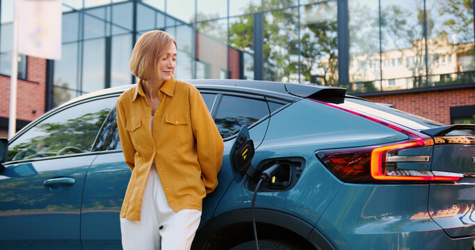 Modern stylish woman connects charging cable parked electric vehicle. Concept of eco transport, green energy, and modern lifestyle. - Powered by Adobe