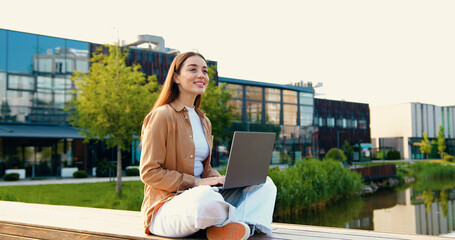 Beautiful and smiling Caucasian girl sitting cross-legged on bench in city park, working remotely on her laptop while enjoying the fresh air and peaceful surroundings