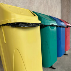 Row of colorful recycling bins used for waste separation in a public area. The image shows yellow, green, blue, and red containers lined up against a concrete wall.