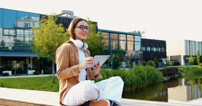 Young woman sitting cross-legged on bench in urban outdoor park, working on a tablet while wearing headphones and eyeglasses, outdoor - Powered by Adobe