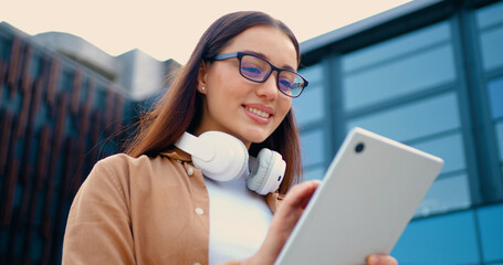 Close-up of beautiful girl smiling while using tablet in urban park during sunny day surrounded by commercial glass buildings in background