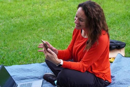 Woman wearing a red sweater, working with a laptop and cell phone in a park - Powered by Adobe