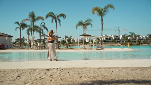Happy young couple in love hugging on sandy beach next to turquoise swimming pool with palm trees, enjoying their summer vacation at luxurious hotel on sunny day - Powered by Adobe