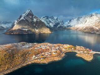 Aerial view of snowy mountains, island with rorbu, sea, bridge, reflection in water, road, cloudy sky at sunset in winter. Damatic landscape. Top drone view of Hamnoy village, Lofoten islands, Norway