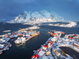 Aerial view of snowy Henningsvaer fishing village, Lofoten islands, Norway in winter at sunset. Top drone view of mountains in snow, sea, boats, bridge, road, cloudy sky, town, rorbuer and houses