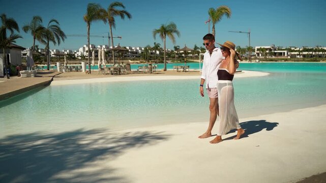 Happy young couple in summer attire walking barefoot along edge of turquoise lagoon at luxury tropical resort, enjoy their romantic vacation under bright sun