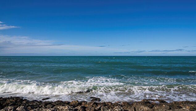 Clear blue sky merges with the ocean, waves softly caress the rocky shore, showcasing nature's splendor