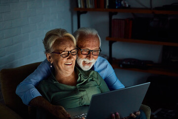 Portrait of an elderly senior happy couple using laptop at night, using modern technology having fun with glowing screen in dark office or at home, watching tv together at home. Shot of a mature activ