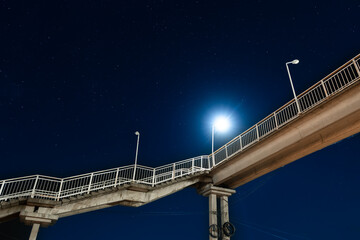 Night view of a pedestrian bridge illuminated by a bright streetlight against a clear starry sky....