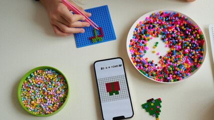 colorful bead art with smartphone guide showing pattern creation for crafting enthusiasts. hand using tweezers and mobile app to follow design template on grid board