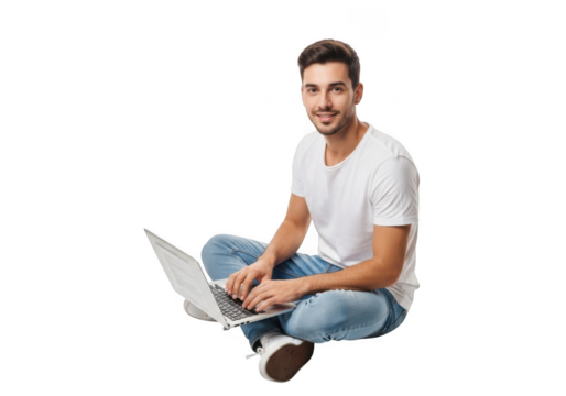 Smiling young man with beard wearing a white t shirt and blue jeans sitting cross legged typing on a laptop computer isolated on transparent background