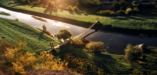 A peaceful river landscape illuminated by warm golden hour sunlight with a small wooden footbridge crossing the water. Green hills, glowing trees and soft atmospheric light create a scenic rural view.