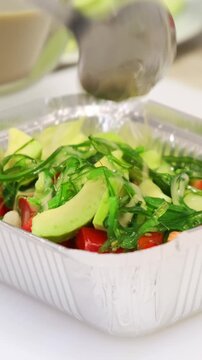 Chef hands, dressing sauce pouring in a vegetable salad with avocado and hiyashi seaweed close up. Healthy and dietary food delivery. Preparing healthy meals in a container box on professional kitchen