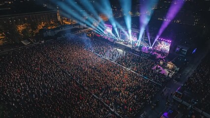 Aerial view of a vibrant outdoor night concert with a massive crowd and dazzling stage lights creating an energetic atmosphere - Powered by Adobe