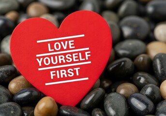 a vibrant red heart featuring the empowering message love yourself first rests among a collection of smooth polished pebbles and stones