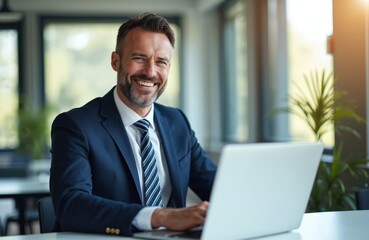 Fototapeta premium Smiling businessman in blue suit works on laptop. Man in office with computer. Professional guy at workplace. Employee with beard uses computer. Man in formal wear sits at desk.