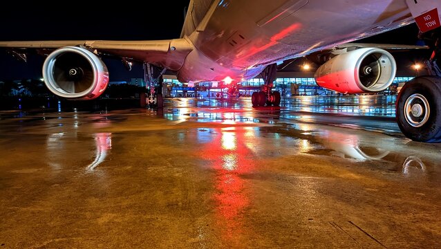 A large commercial aircraft parked on wet apron of Soekarno&ndash;Hatta International Airport at night, captured from a low angle showcasing its powerful engines, landing gear, and reflective ground lights